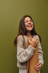 Cheerful and stylish preteen girl in modern knitted sweater and autumn outfit holding rolled paper and laughing while standing isolated on green, girl radiating autumn vibes concept