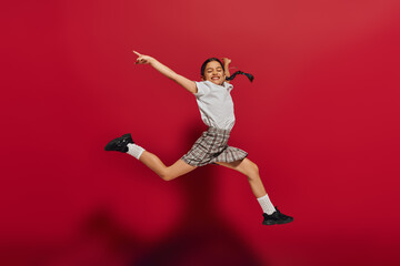 Fototapeta na wymiar Full length of pleased and stylish preteen girl with hairstyle wearing t-shirt and plaid skirt while jumping and having fun on red background, hairstyle and trendy accessories concept