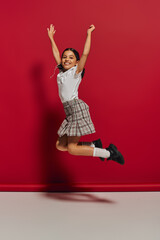 Positive and trendy brunette preteen girl in t-shirt and checkered skirt jumping while having fun and looking at camera on red background, hairstyle and trendy accessories concept