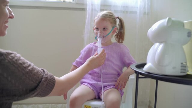 Cute little little girl inhaling mist from nebulizer at home , mom helping and entertain her 