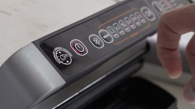 A man's finger presses the power button on a home electric grill to turn it on. Household electrical appliances in the home kitchen. Black electric grill control panel close-up.