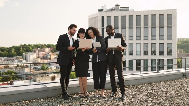 Diverse team of foreign investors standing on flat rooftop with personal gadgets and discussing marketing strategy of new business project. Officially dressed colleagues watching online presentation.
