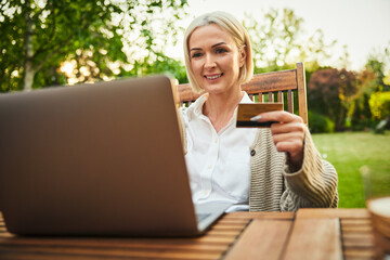 Mature woman paying for online shopping on laptop with credit card while sitting by garden table...