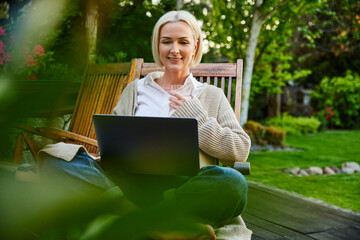 Adult woman sitting relaxed with laptop on garden terrace during summertime watching looking at...