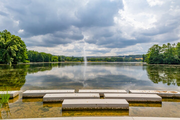 Fototapeta premium Panoramic landscape of Lake Echternach with waterjet from a fountain, green leafy trees on background, concrete platforms on shore, reflection on water surface, cloudy summer day in Luxembourg