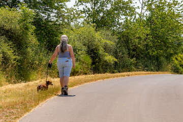 Obraz premium Female adult hiker walking with her dog on left side of a country road, wild vegetation and green trees in background, cap, casual clothing, shorts, tank top, sunny summer day in Utscheid, Germany