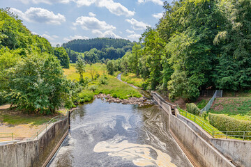 Water flowing from Staumauer Bitburger dam into Prum river, valley and mountains with leafy trees...