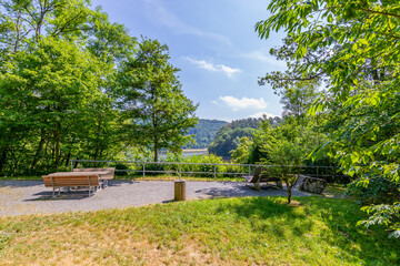 Panoramic viewpoint surrounded by abundant green vegetation and leafy trees with Stausee Bitburg reservoir lake in background, benches and wooden table, sunny day in Germany