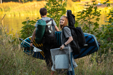 Back view of happy young couple going home after camping together with backpacks and paddleboard. Woman turned round and looking at camera with smile. One shot in series of camping 