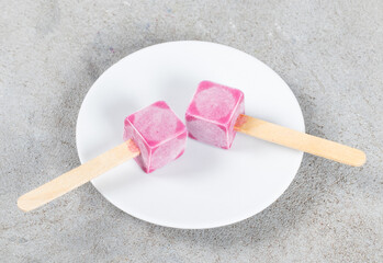 Two black currant ice creams in the shape of small cubes on a wooden stick. On a plate. Top view