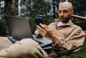 A man working on a laptop and a smartphone is lying in a hammock in the backyard