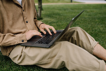 Concept photo of a young man in casual clothes using a laptop for remote work while sitting on a green lawn