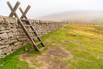 Mourn Wall with ladder on the back of Slieve Donard mountain with foggy sky, white clouds. Mourne Mountains range in Northern Ireland.