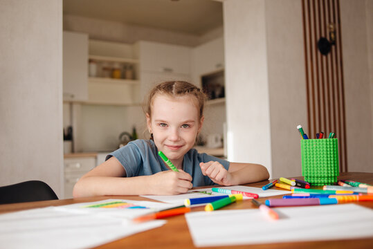 A girl draws at home with felt-tip pens on white paper on the background of the kitchen