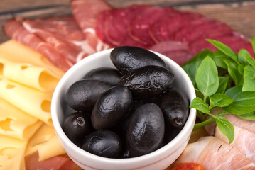 Selective focus on olives against the background of cuts of prosciutto, salami, bacon, cheese and green basil.