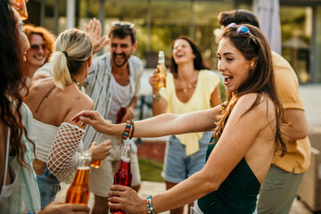 Group of friends dancing and smiling gather on the poolside, toasting with their cocktails to the beautiful colors painting the sky.