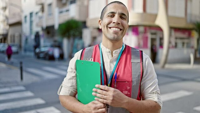 Young hispanic man volunteer smiling holding clipboard at street