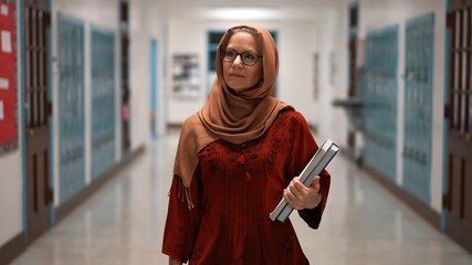 Front view closeup of happy smiling muslim teacher wearing headscarf walking down a hallway in an empty school holding books. - obrazy, fototapety, plakaty