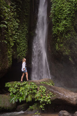 Naklejka na ściany i meble Waterfall in tropical jungle and alone woman tourist. Leke Leke waterfall in Bali, Indonesia.