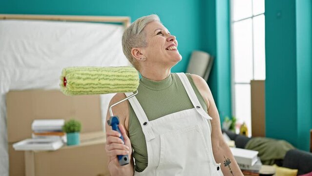 Middle age grey-haired woman smiling confident holding paint roller looking around at new home