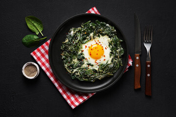 Fried eggs with fresh spinach and creamcheese on a black plate on the red napkin on the dark background, top view