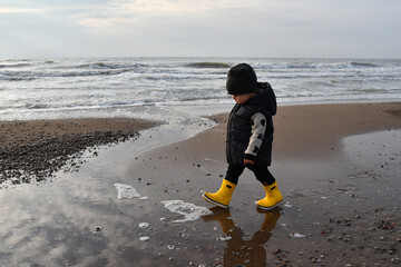 Little Toddler boy wearing yellow rubber boots walking down the sea in autumn season - obrazy, fototapety, plakaty