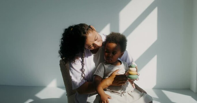 Mom with a child of African-American appearance, dressed in white clothes, playing pyramid while sitting in a chair in a white room