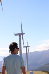 Man with blue t-shirt and sunglasses observing the landscape on top of a mountain with windmills in the background, in Monte Pindo, Carnota, Galicia, Spain. Upright image.
