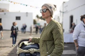 Keuken foto achterwand Canarische Eilanden Portrait of young stylish woman wearing green padded jacket, hoodie, wool cap and sunglasses pershing stroller down cobled street of touristic town of Tequise, Lanyarote, Spain