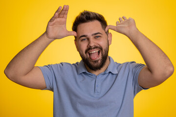 Comical funny young caucasian man making playful silly facial expressions and grimacing, fooling around, showing tongue. Handsome crazy adult guy bullying isolated on studio yellow background indoors