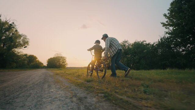 Happy grandfather teaches his grandson to ride a bicycle on a road at sunset