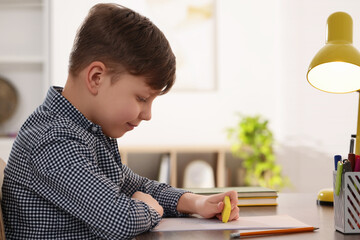 Little boy erasing mistake in his notebook at wooden desk indoors