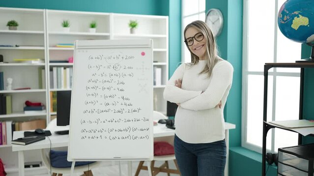 Young pregnant woman teacher teaching maths lesson standing with arms crossed gesture at classroom
