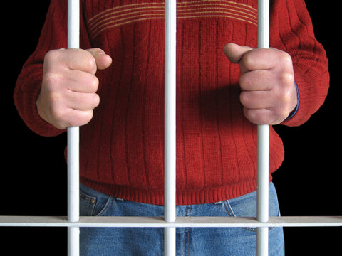 Prisoner wearing a red sweater holding iron bars behind bars, concept of crime, imprisonment, and criminal justice