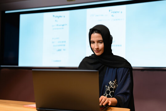 Teacher or Student concept of woman wearing Abaya Hijab with projector screen as background. Modern classroom using digital screen with instructor or Arab Student mock up. Female Emir using laptop