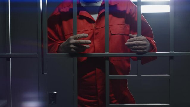 Elderly prisoner in orange uniform holds metal bars, stands in prison cell. Guilty criminal or thief serves imprisonment term for crime. Inmate in jail, detention center or correctional facility.