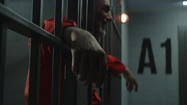 Elderly prisoner in orange uniform cries, holds his hands on metal bars. Criminal serves term of imprisonment in prison cell. Depressed inmate stands and walks behind bars in jail or detention center.