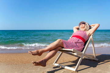Smiling elderly woman is sitting in a sun lounger on a sandy beach near the sea on a sunny day. The lady enjoys nature and relaxation. Active lifestyle and health. Space for text.