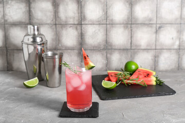 Glass of tasty watermelon cocktail with shaker and lime on grunge grey table near wall