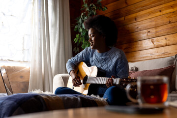 Happy african american woman sitting on sofa playing guitar and using laptop in sunny living room