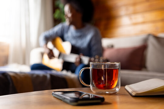 Notebook, tea and phone on table and african american woman playing guitar in background, copy space