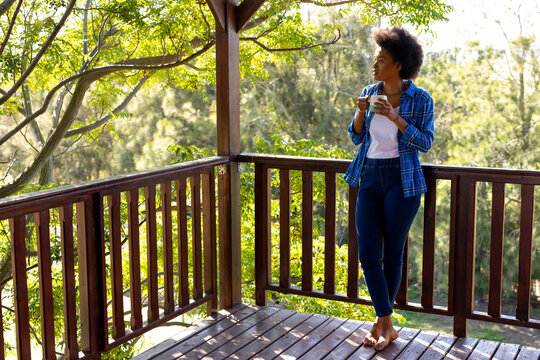 Thoughtful african american woman standing on balcony with cup of tea, looking at trees, copy space