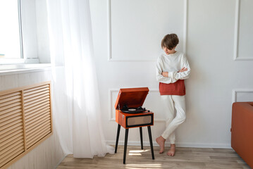 Teenager young boy standing near retro wooden vinyl turntable, looking on player and listening music indoor, in a white minimalist room interior