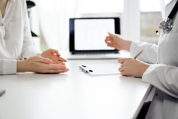 Doctor and patient sitting and discussing something near each other at the white desk in clinic. Female physician is pointing into laptop screen. Medicine concept