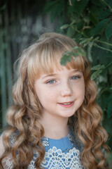 Fototapeta premium Portrait of a girl with long blonde hair on the street. A 9-11-year-old girl against a background of green plants, smiling and looking at the camera
