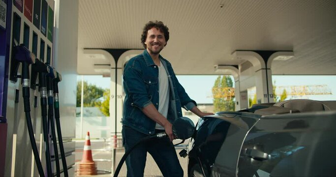 Portrait of a curly-haired brunette guy who refuels a gray convertible. Happy car ride, gas station stop