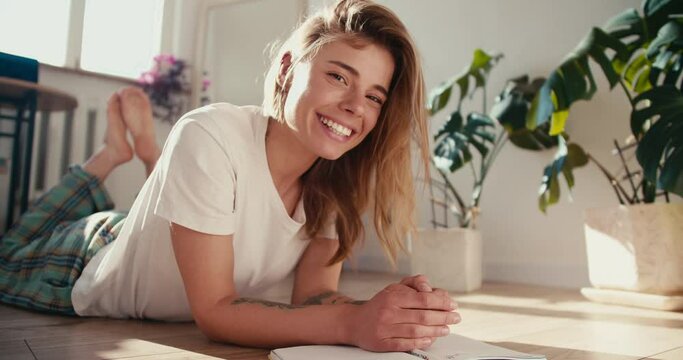 a blonde girl in a white T-shirt lies on the floor next to her are houseplants in a sunny room. Portrait of a beautiful smiling girl who lies on the floor in the morning