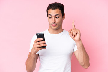 Young caucasian man isolated on pink background using mobile phone and lifting finger