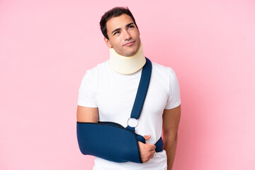 Young caucasian man with broken arm and wearing a sling isolated on pink background and looking up