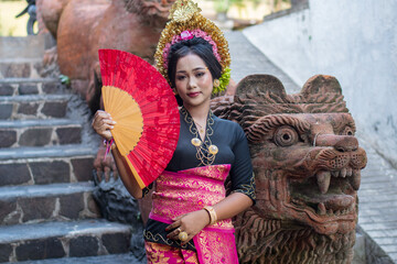 Indonesian girl with traditional black-red costume and crown in bali relief wall and a statue of a...